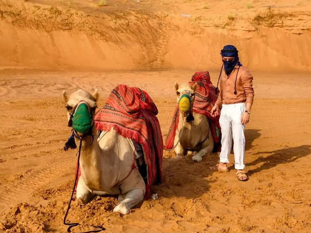 Camel handler with two resting camels on the red sand at the desert camp